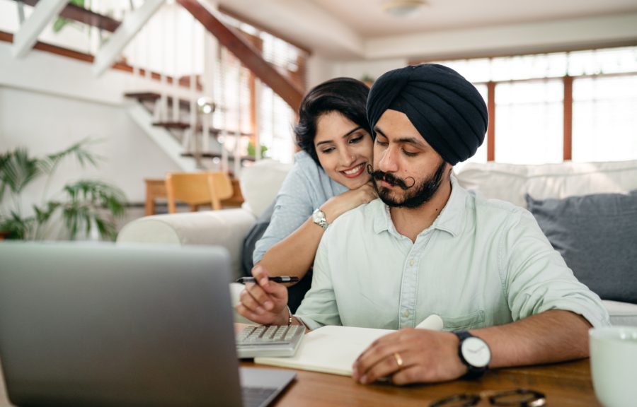 Serious Young Sikh Man With Hugging Wife Counting On Calculator At Home