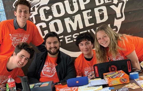 Global Improvisation Initiative Members At A Stall For One Of Their Comedy Events Behind A Branded Banner Wearing Branded Merch